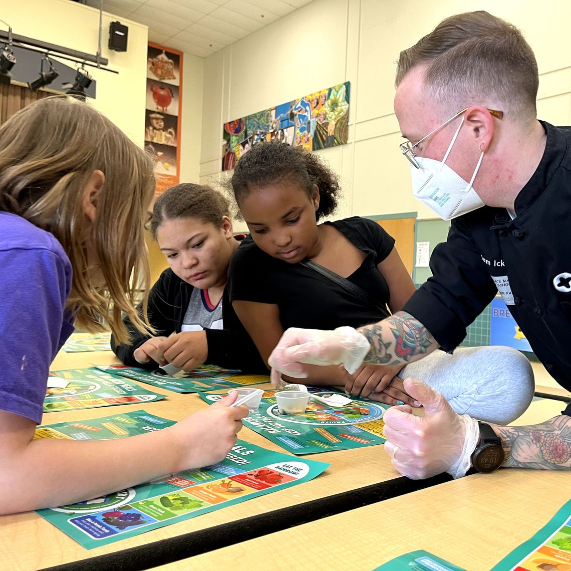 Chef Sam of Project Bread is at a table in the cafeteria talking to students about a sample of a new recipe they just tried