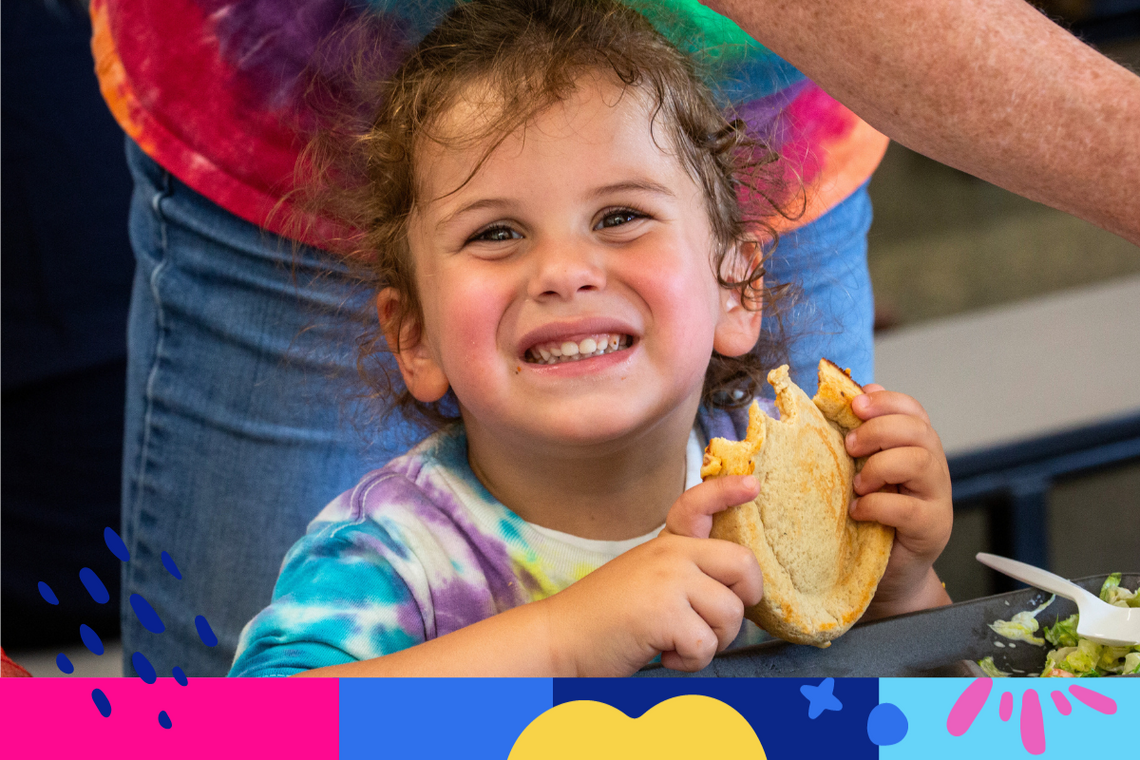 A little girl smiles big holding a sandwich at a Summer Eats program