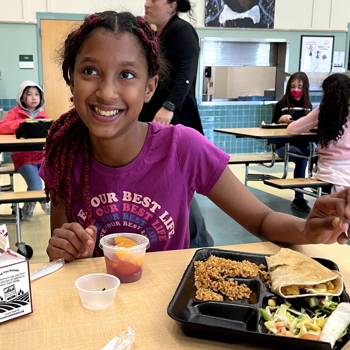 A young student with braided hair sits at cafeteria table in front of a lunch of beans and rice, fresh fruit and milk